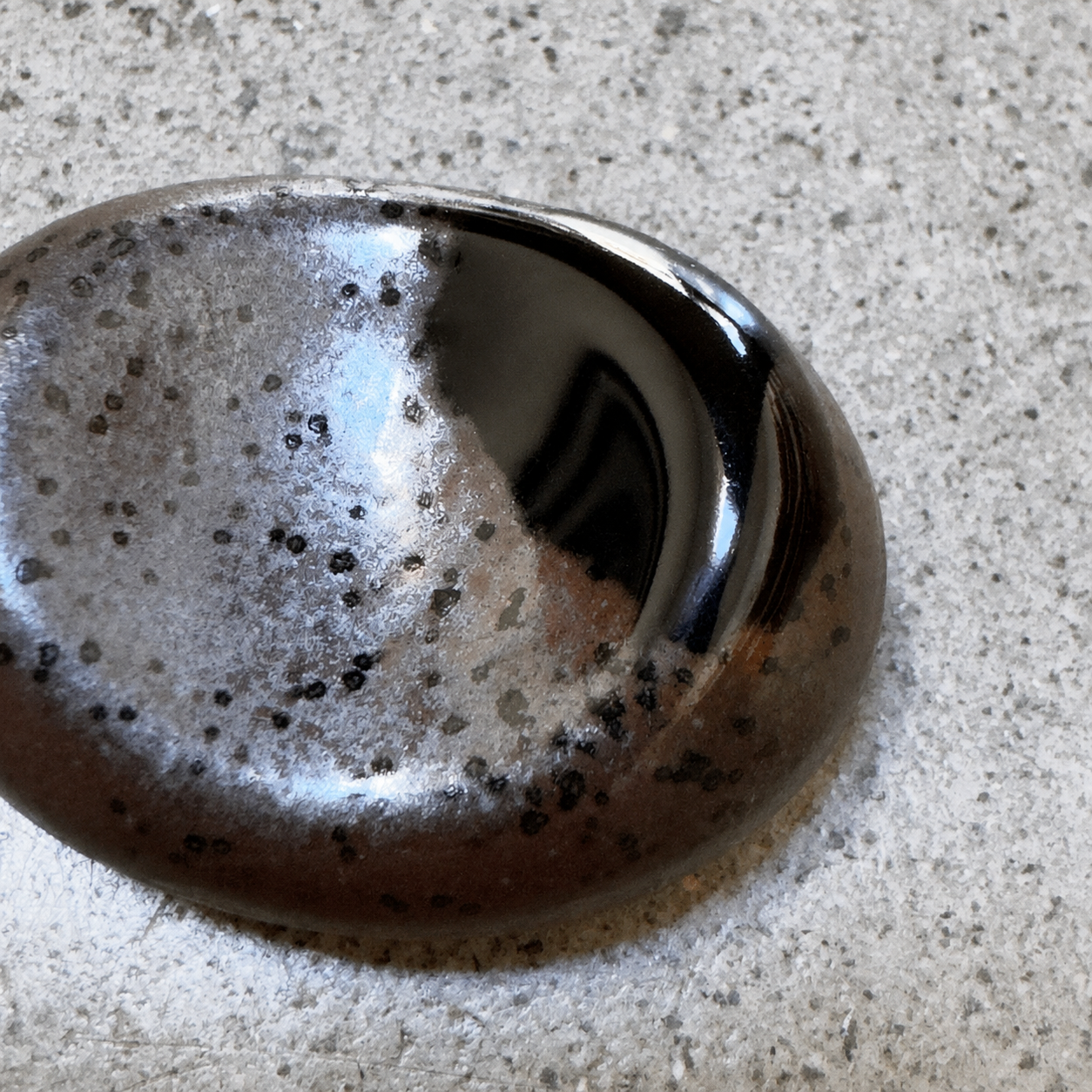 A polished dark brown and black Japanese ceramic chopstick rest with black speckles, resting on a textured light grey concrete surface.