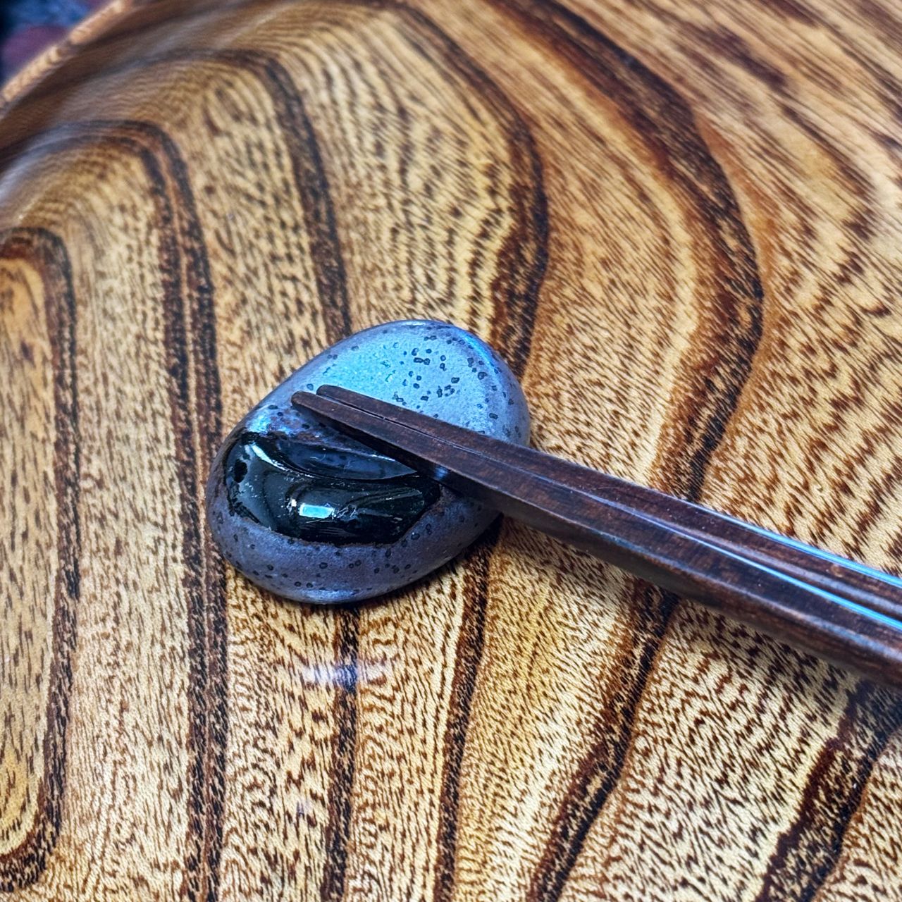 A black Japanese ceramic chopstick rest with dark speckles, held by dark brown chopsticks on a highly polished wood surface with dramatic swirling grain.