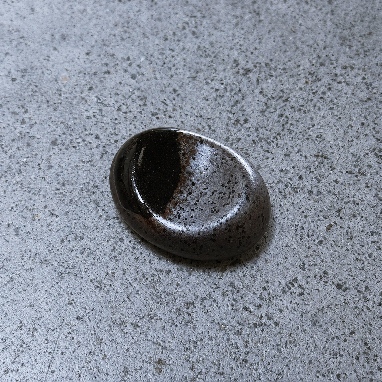 A rounded, polished Japanese ceramic chopstick rest with a dark brown and black speckled glaze, resting on a textured grey concrete surface.