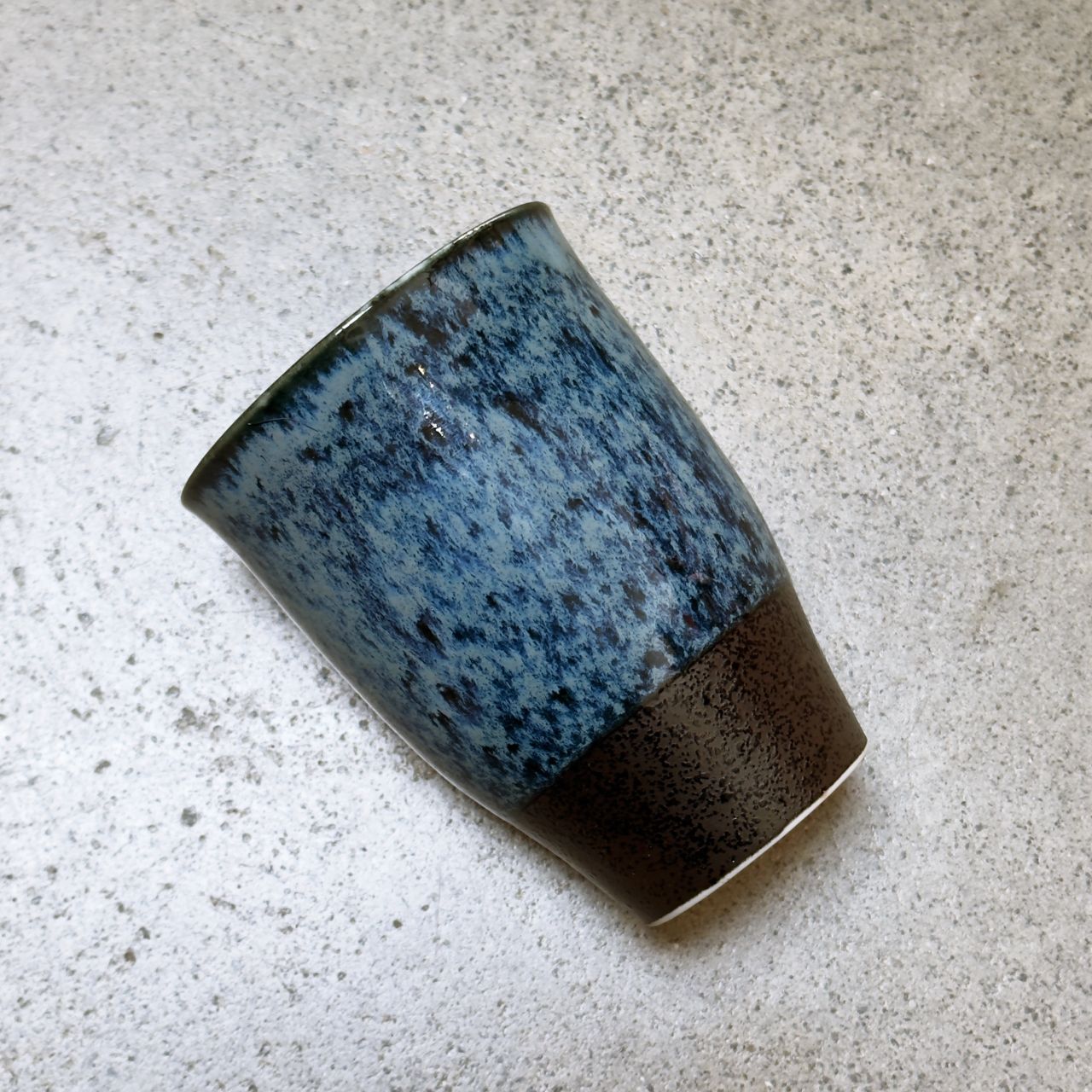 A handmade ceramic cup with a textured blue speckled glaze on the upper half and a dark brown glaze band at the base, resting on a light grey concrete surface.