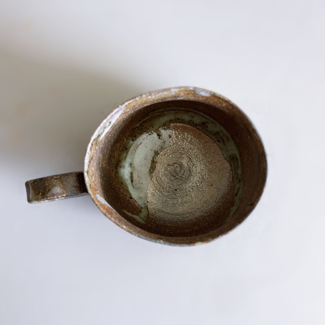 Top-down view of a handmade ceramic mug with a rustic, earthy brown and beige glaze and a handle, set against a plain light background.