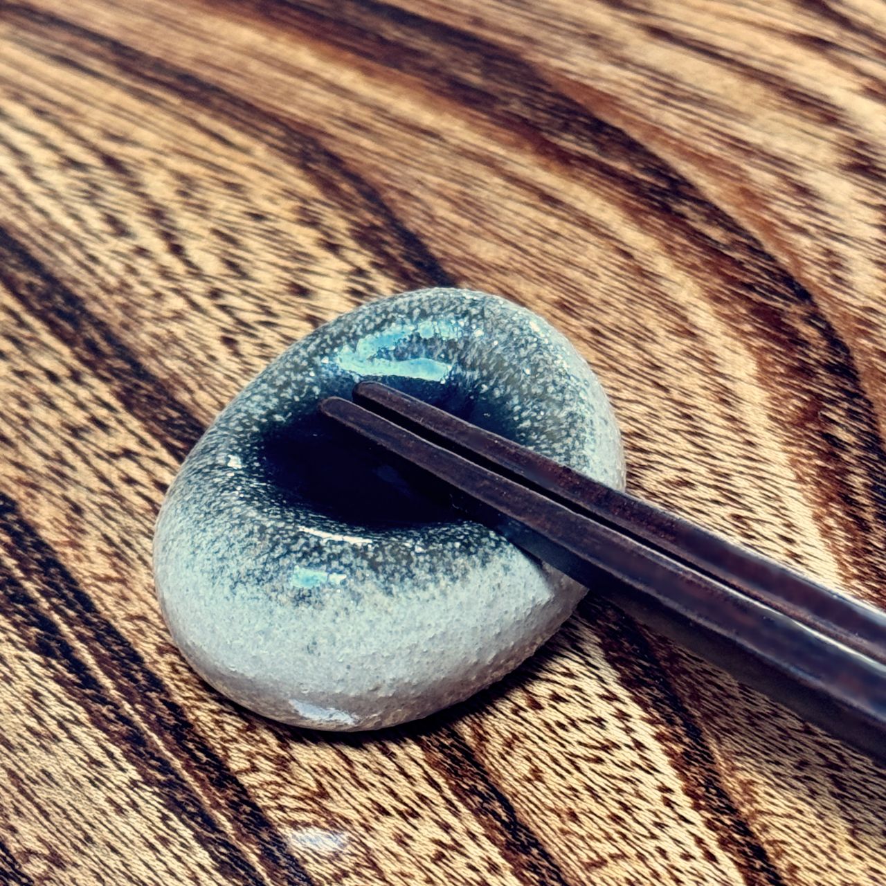 An oval Japanese ceramic chopstick rest with a dark glazed centre and speckled light grey exterior, shown on a wooden surface with chopsticks resting across the rim.