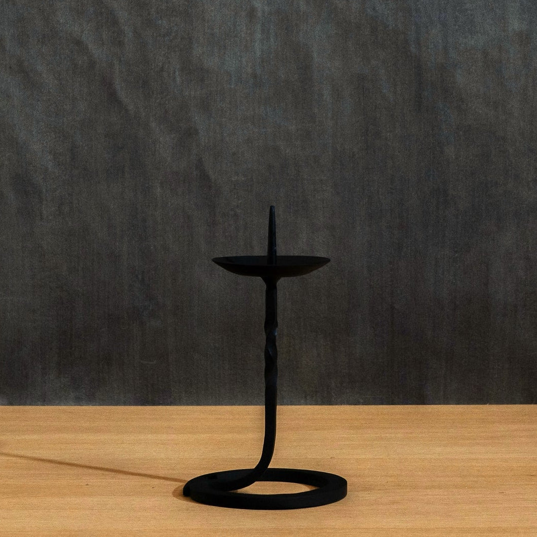 A black Japanese handcrafted iron candle holder with a twisted stem and circular base, standing on a light wood surface against a dark textured wall.