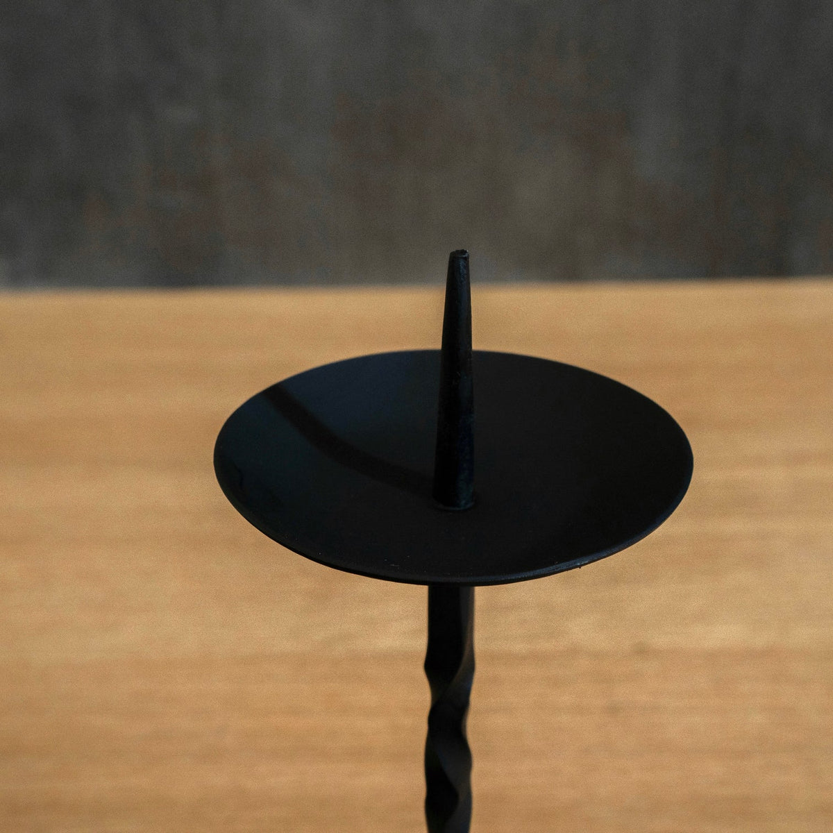 A black Japanese iron candle holder with a flat circular top and a twisted stem, shown on a wooden surface against a dark grey background.