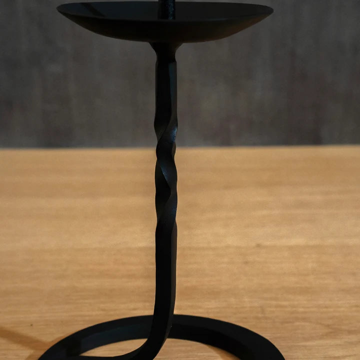 A black Japanese iron candle holder with a twisted stem and circular base, shown on a wooden tabletop against a dark grey wall.