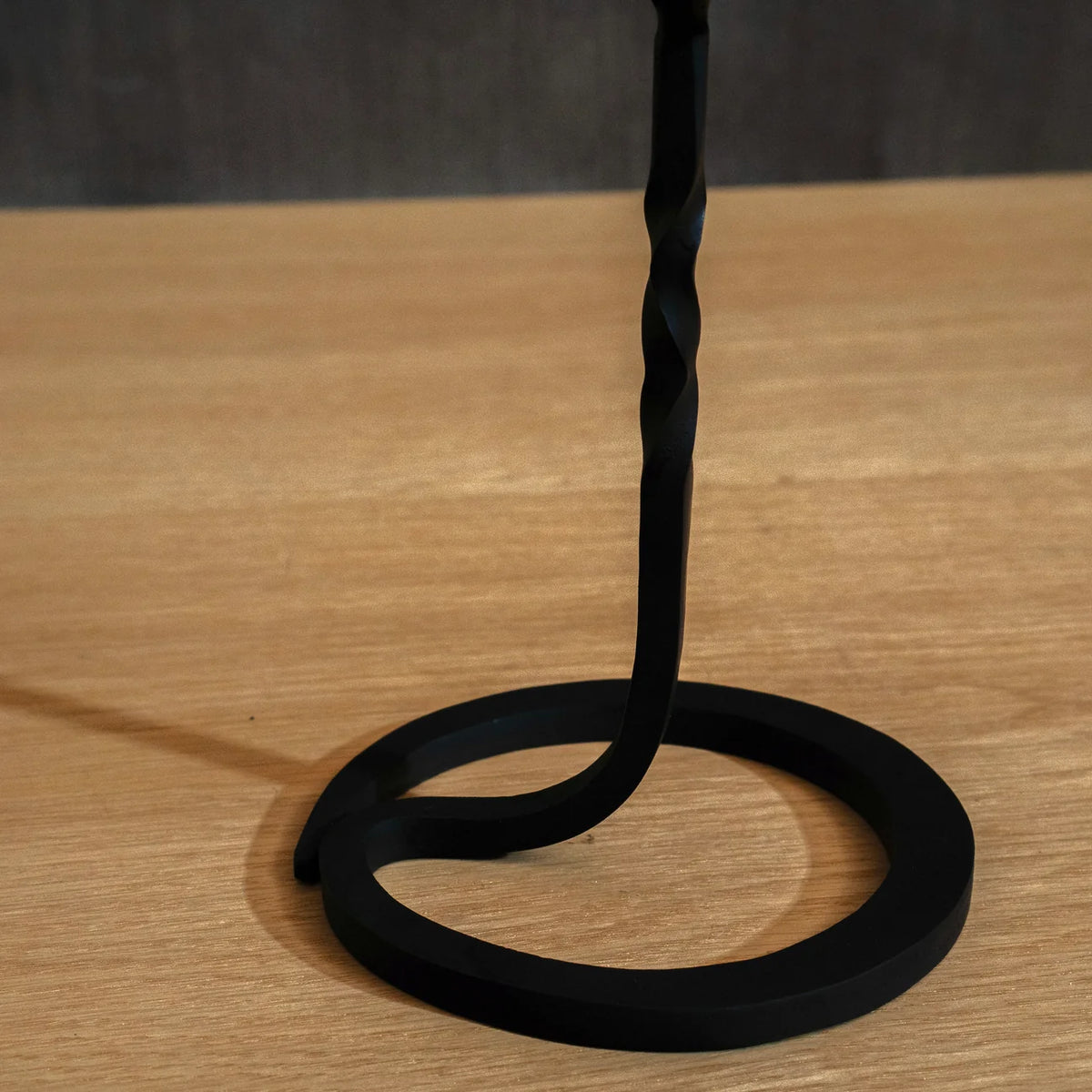 A black Japanese iron candle stand with a twisted vertical stem and a circular base, photographed on a light wood tabletop against a dark grey background.