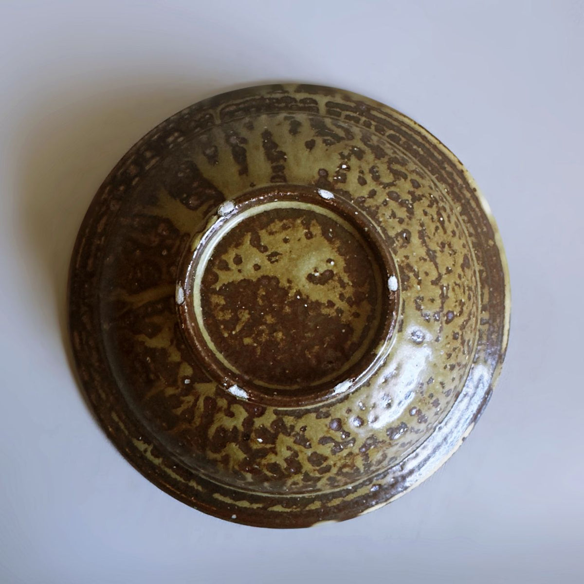 A top-down view of a ceramic bowl with a textured, speckled glaze in earthy brown and olive green tones, set against a plain light gray background.