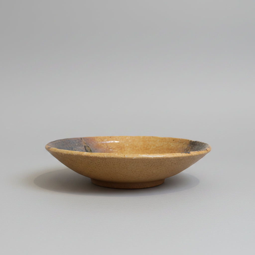 A handmade Japanese ceramic shallow bowl with a speckled brown exterior and a dark grey glazed interior, sitting on a grey background.