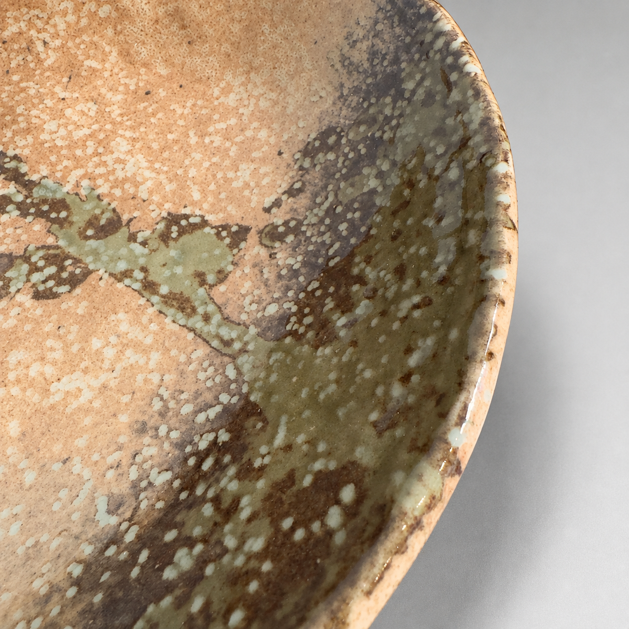 Close-up of a Japanese handcrafted ceramic bowl showing a textured glaze with mottled brown, and muted green tones on a light grey background.