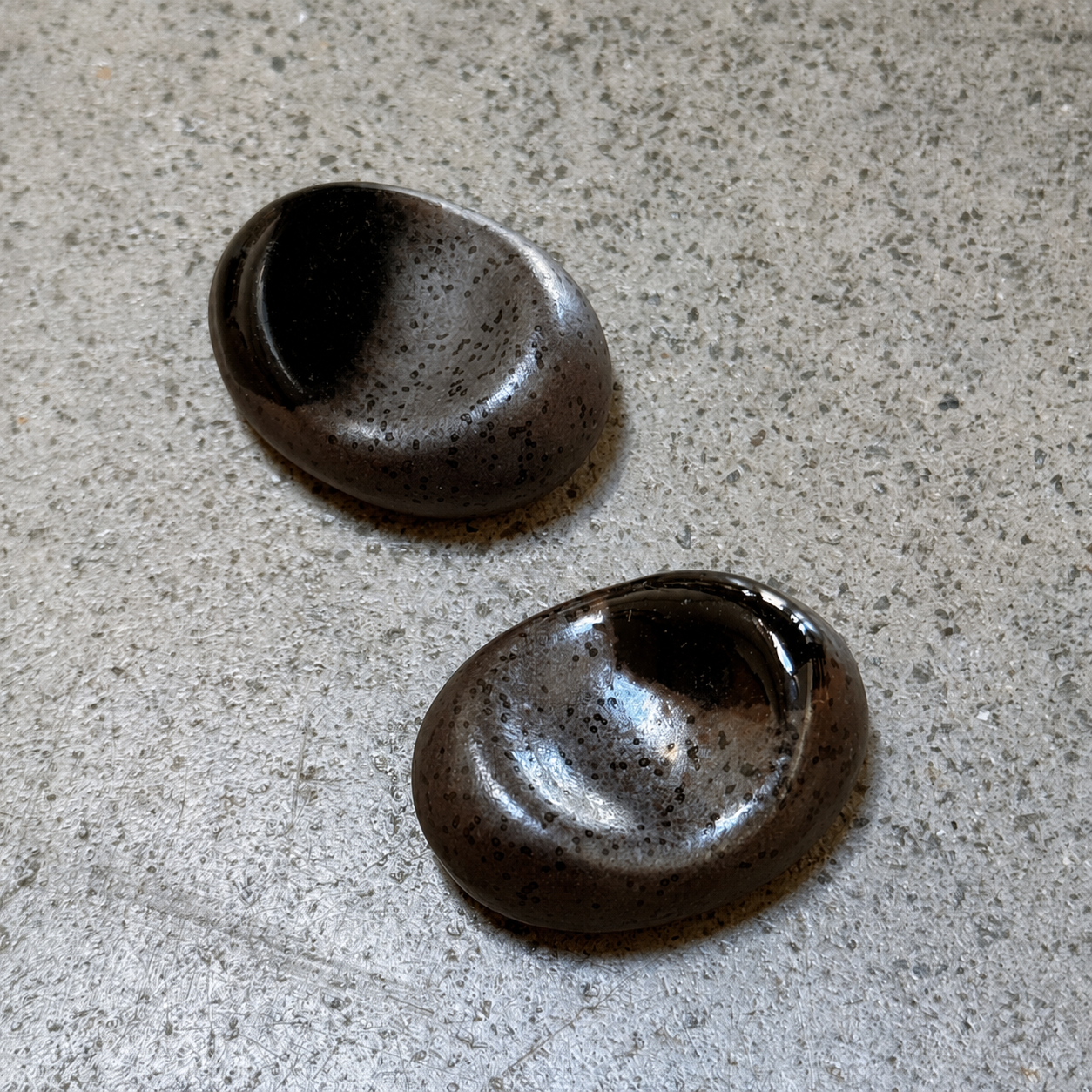 Two oval Japanese ceramic chopstick rests with a  dark brown and black speckled glaze, resting on a textured grey surface.