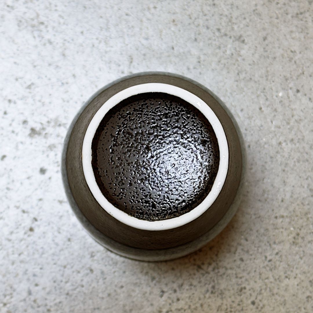 A top-down view of a bottom of round ceramic cup with a textured dark brown glaze set on a speckled light grey concrete surface.