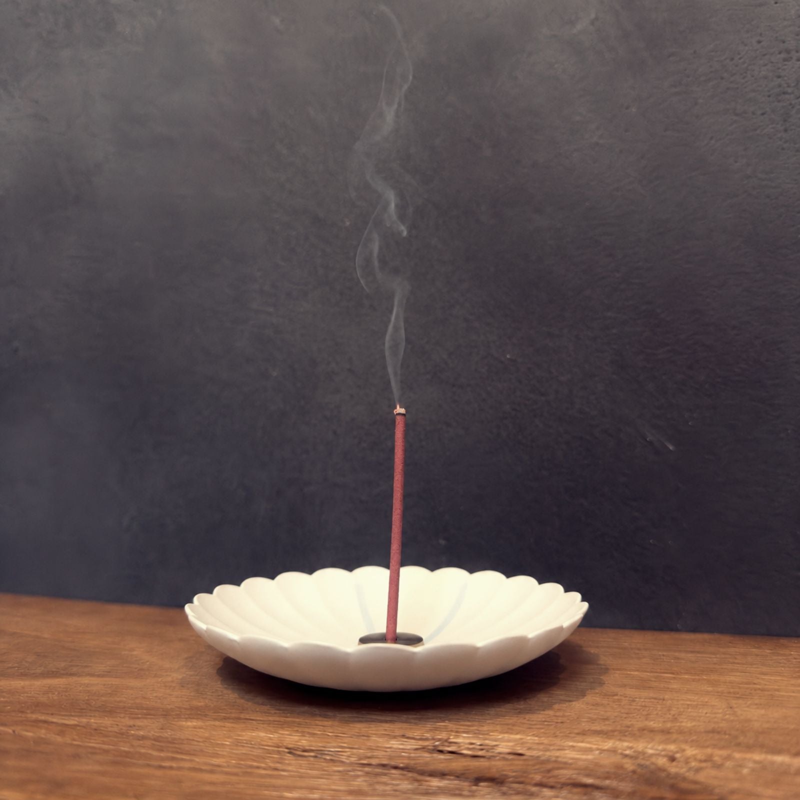 Lit Japanese incense stick with rising smoke in a white scalloped bowl on a wooden bench, from Senju Living.