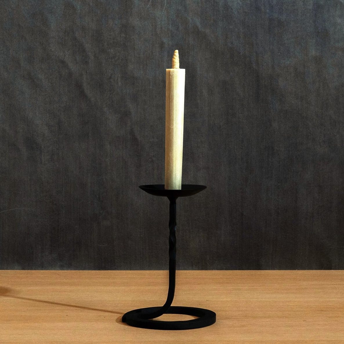 A black Japanese hand forged  candle holder with a twisted stem and circular base, holding a single Japanese warosoku candle, set on a light wood surface against a dark textured wall.