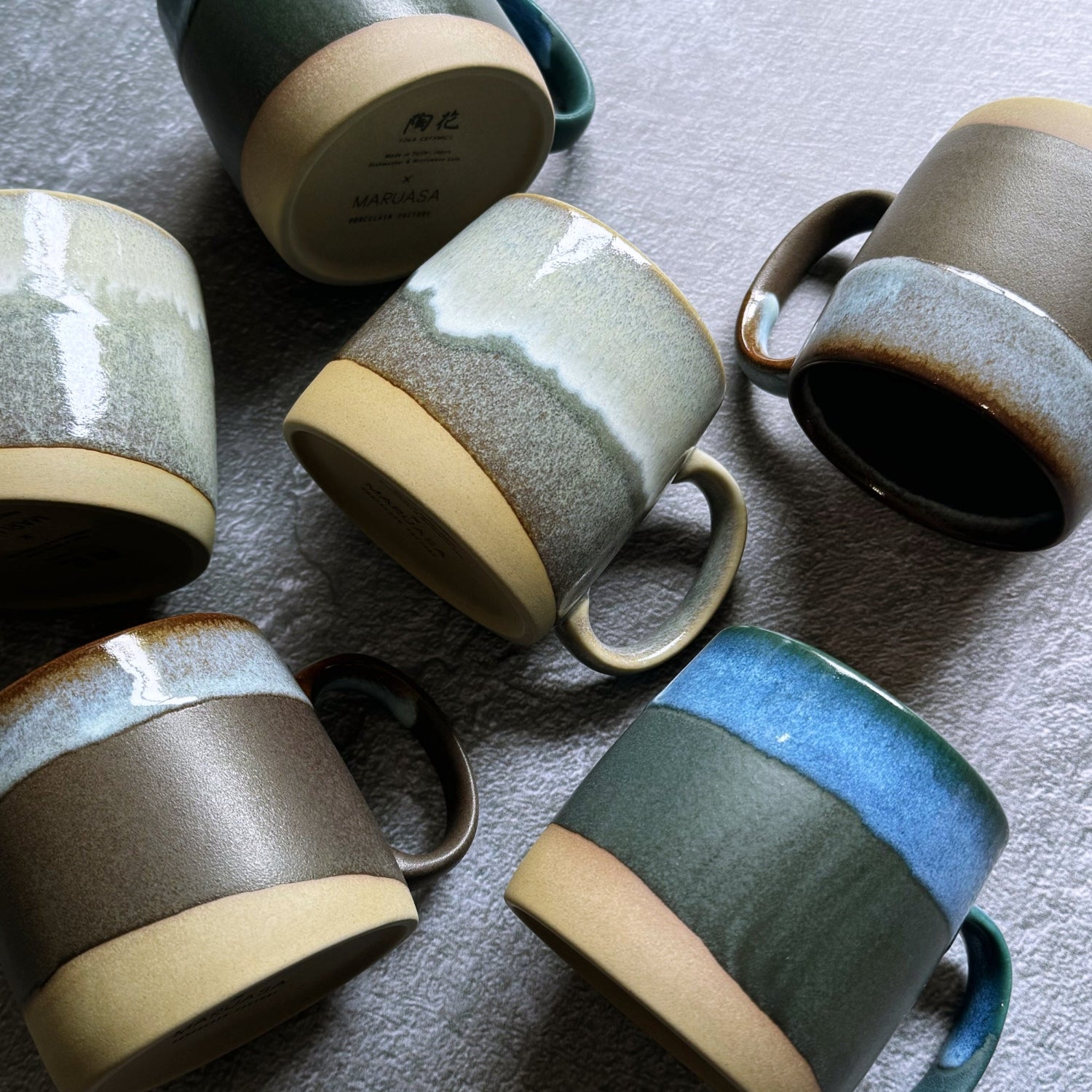 Flat lay of TOKA Japanese ceramic mugs in various glazes, showing the Maruasa Porcelain Factory maker stamp