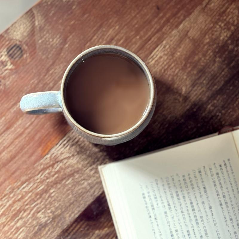 Top-down view of a TOKA Japanese ceramic mug with coffee on a rustic wood table next to an open book, quiet daily ritual.