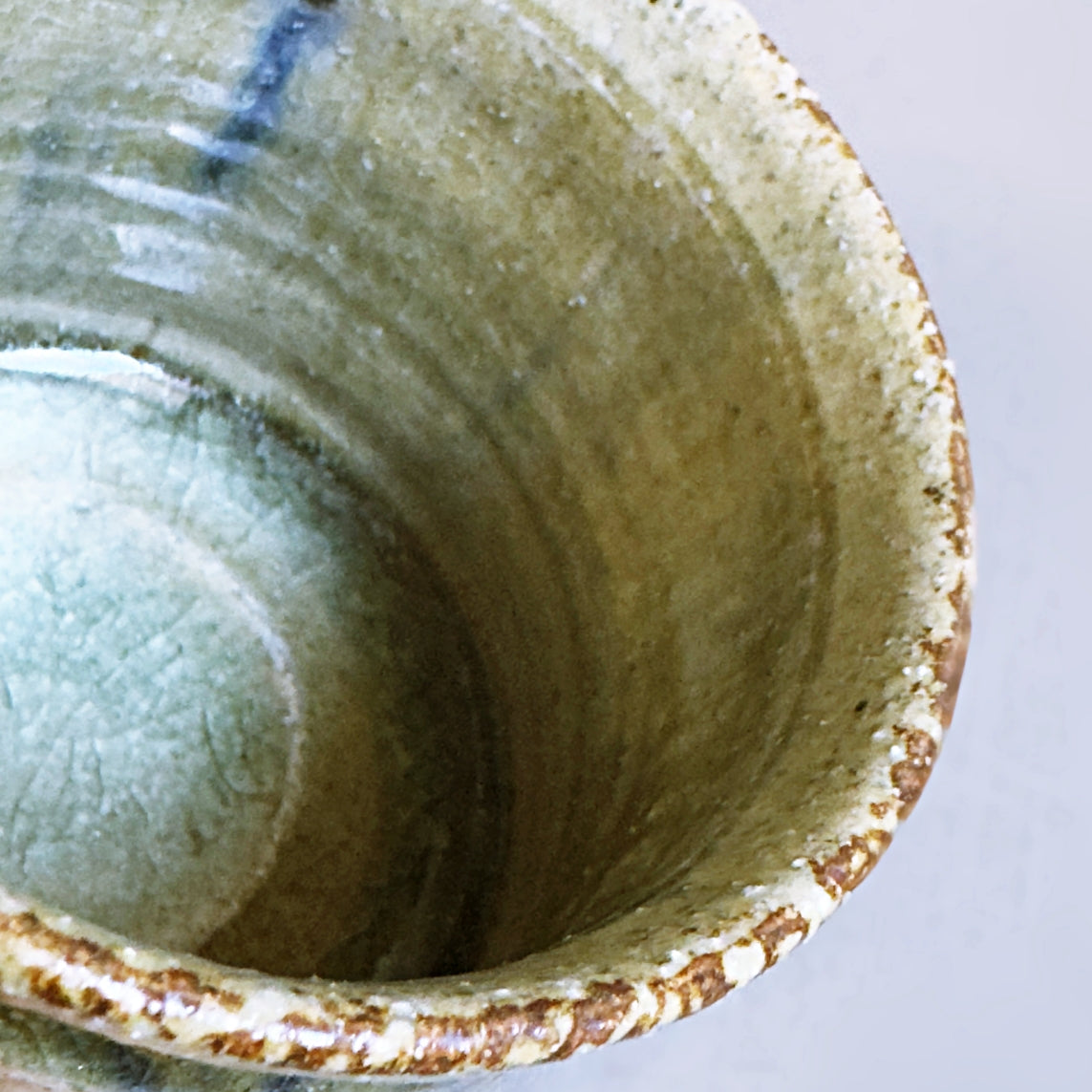 Close-up of a handmade Japanese ceramic cup showing the textured interior glaze with a light green and beige finish and a rustic brown speckled rim on a light grey background.