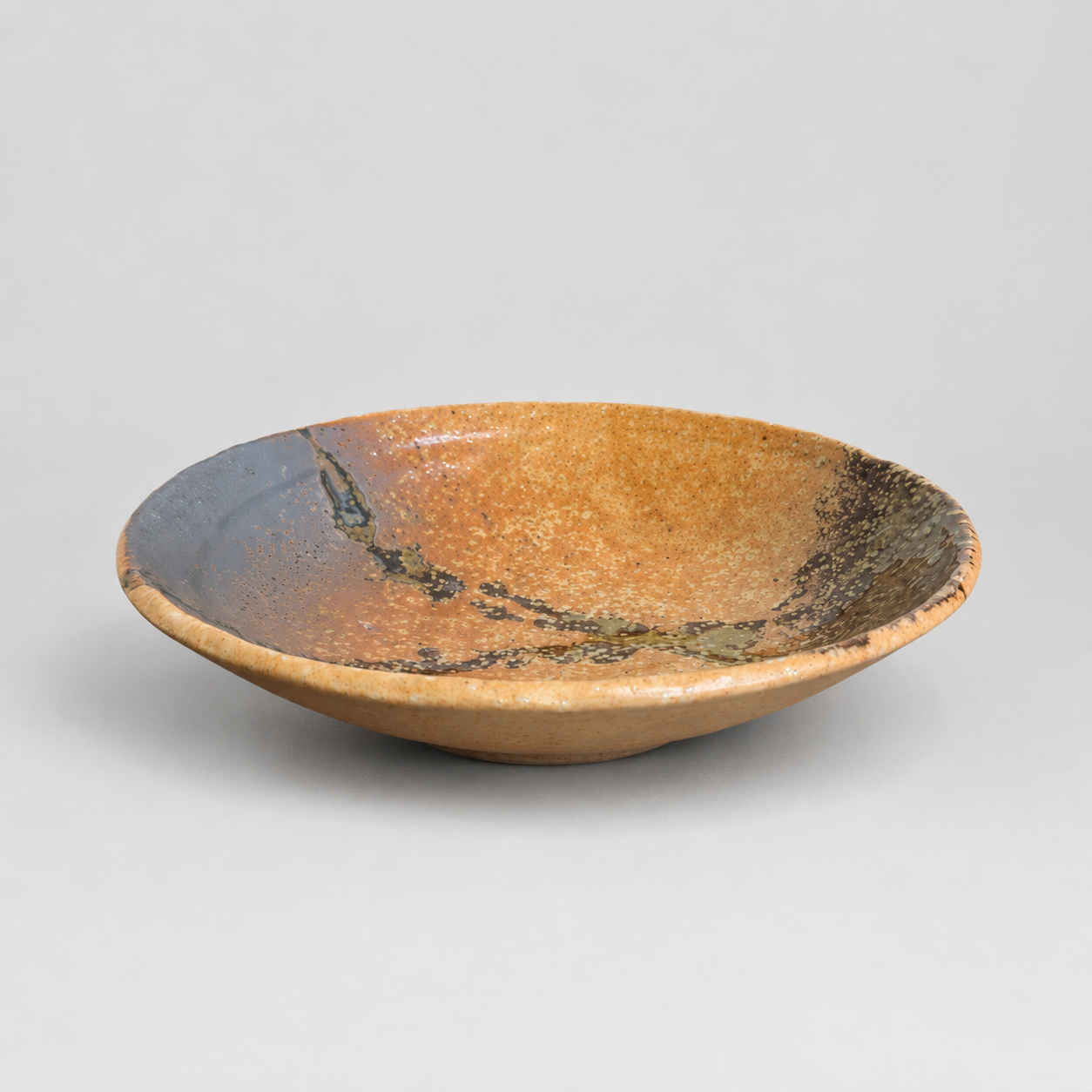 A shallow ceramic bowl with a textured, earthy glaze featuring a gradient from dark grey to warm ochre brown, with dark green and black mineral-like speckles, set against a neutral light grey background.