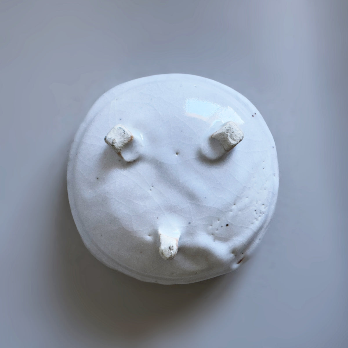 A bottom of a roundish white ceramic bowl with three small raised feet, photographed from above on a plain light grey background.
