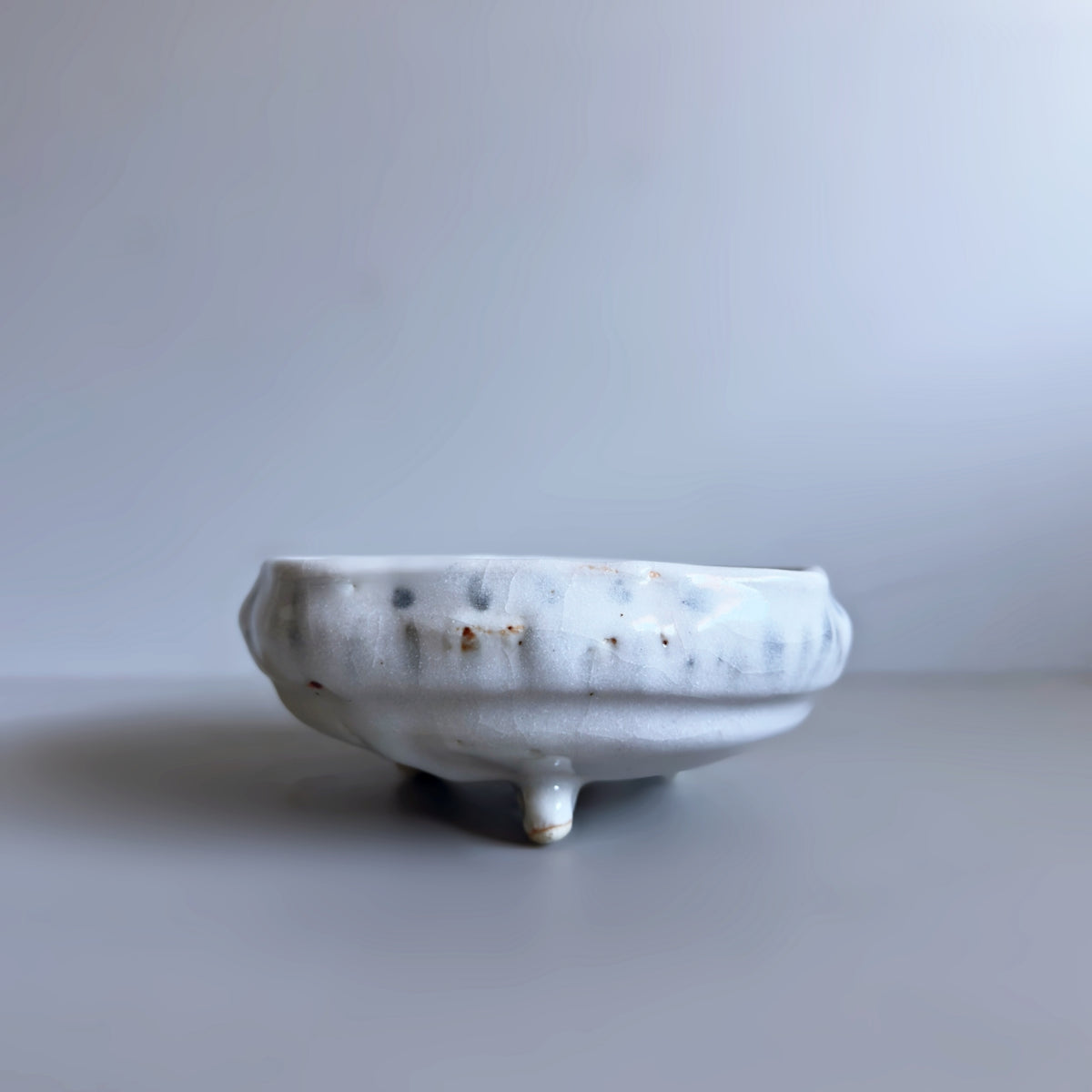 A Japanese handmade ceramic bowl with a textured, ribbed exterior and three short legs, finished in a glossy off-white glaze with subtle speckles and brownish spots, set against a neutral grey background.